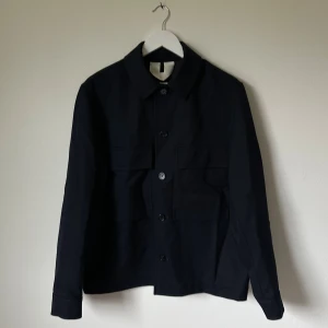 Mörkblå overshirt med fickor, strl 50 - Stilren mörkblå skjortjacka/overshirt i storlek 50 med klassisk krage och stora frontfickor med lock. Jackan har knäppning med knappar hela vägen och långa ärmar med knapp vid mudden. Perfekt för dig som gillar minimalistisk och clean look. 4D18