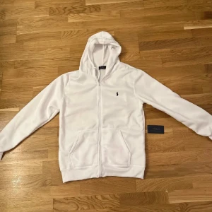 Vit polo zip - Säljer en stilren vit hoodie från Polo Ralph Lauren med dragkedja och det klassiska logotypbroderiet på bröstet. Perfekt för en avslappnad look.