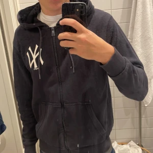 Svart New Era Yankees hoodie XS - Svart hoodie från New Era med New York Yankees-logga i vitt på bröstet. Tröjan har dragkedja, huva med snörning och två fickor framtill.