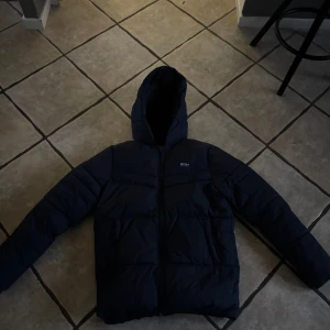 Mörkblå pufferjacka från BOSS - Säljer en mörkblå pufferjacka från BOSS med huva och dragkedja framtill. Jackan har ett stilrent, quiltat mönster och långärmade ärmar. Perfekt för kalla dagar och har en snygg, modern look som passar till streetwear.