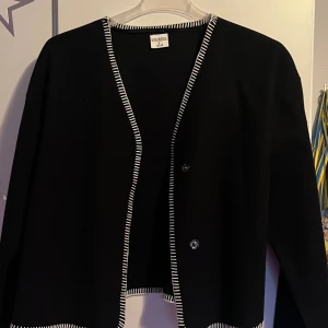 Svart cardigan jacka från Stories - Svart cardigan jacka från Stories i storlek S. Jackan har vita dekorativa sömmar längs kanterna och nederdelen, samt två knappar framtill. Stilren och enkel design med lång ärm och rak passform.