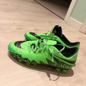 Nike Hypervenom gröna fotbollsskor - Säljer ett par Nike Hypervenom fotbollsskor i en riktigt grön färg med svarta detaljer och grafiskt mönster längs sidorna. Skorna har snörning och platt sula med dobbar för gräsplan. Snygg, sportig design som sticker ut på planen.