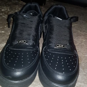 Nike Air Force 1 helsvarta sneakers - Säljer ett par klassiska Nike Air Force 1 sneakers i helsvart utförande. Skorna har en låg siluett, snörning och perforerad tå för extra ventilation. Tillverkade i skinn med platt sula och diskret Nike-logga på sidan. Perfekt för dig som gillar stilrena och tidlösa sneakers.