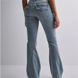 True Religion Joey Bootcut Jeans - Snygga ljusblå jeans från True Religion, modell Joey med bootcut passform. Jeansen har ikoniska fickor med dekorativa sömmar och silverfärgade knappar. Storlek 29, säljs inte längre så går inte får ta i. Lågmidjade byxor.