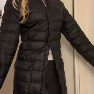 Svart lång pufferjacka från H&M - Säljer en svart, lång dunjacka från H&M i storlek 32. Jackan har stora fickor med knappar framtill. Perfekt för kalla dagar med sin quiltade design och långa ärmar.