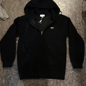 Svart hoodie från Lacoste med dragkedja - Svart hoodie från Lacoste med klassisk krokodillogga på bröstet. Tröjan har huva med snörning, praktisk dragkedja och två fickor framtill. Tillverkad i mjuk bomull, perfekt för chill dagar. Långärmad och normal passform.