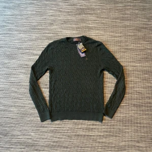 Grön kabelstickad tröja Morris M med tags - Kabelstickad mörkgrön tröja från Morris i 100% merinoull. Rund hals och ribbade muddar vid ärmslut och nederkant. Perfekt för dig som gillar klassisk stil med snygga detaljer. Diskret Morris-logga på bröstet. Bara använd nån enstaka gång. OBS tagsen sitter inte på men är avklippta. Nypris: 1599kr
