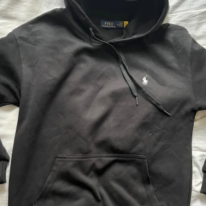 Svart hoodie från Polo Ralph Lauren - Hej!  Säljer en Svart hoodie från Polo Ralph Lauren med klassisk vit broderad logga på bröstet. Tillverkad i mjuk bomull med huva och snörning samt en stor magficka framtill. En stilren och tidlös tröja med riktigt premiumkänsla.  Tröjan är märkt som storlek M men passar bäst för dig som vanligtvis använder storlek S.  Vill du veta något mer eller vill ha fler bilder så är det bara att skriva! Priset är såklart inte hugget i sten!
