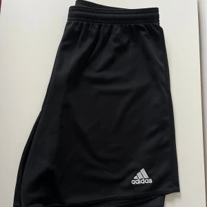 Svarta sportshorts från Adidas - Svarta sportiga shorts från Adidas med klassisk logga broderad i vitt på ena benet. De har elastisk midja och är tillverkade i ett lätt och svalt polyestermaterial, perfekta för träning eller sportiga dagar.