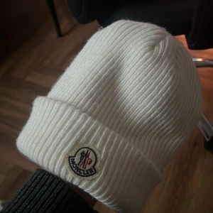 Vit ribbstickad mössa från Moncler - Säljer en snygg vit ribbstickad mössa från Moncler med klassisk uppvikt kant och broderad logotyp framtill. Mössan är tillverkad i mjukt material och har en stilren look som passar perfekt till kyliga dagar.