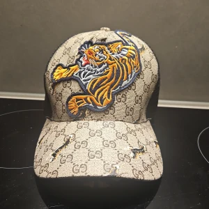 keps med tiger och GG-mönster - Snygg keps med klassiskt GG-mönster i beige och brunt, broderad tiger framtill och orangea XXV-detaljer bak. Baksidan är i svart mesh med justerbart spänne i läder. Perfekt statement-piece för dig som vill sticka ut.
