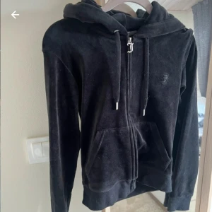 Svart velour hoodie med dragkedja - Mysig svart hoodie i mjuk velour med huva och snörning. Tröjan har dragkedja framtill och två fickor, samt diskret broderad logga på bröstet. Perfekt för chill dagar eller när du vill vara extra bekväm.