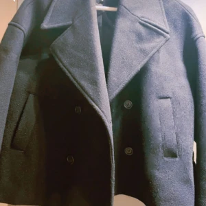 Svart skepparkavaj i ull, storlek M köpt 2025 - Säljer en svart skepparkavaj i ull med klassisk dubbelknäppning och breda slag. Jackan har två raka fickor framtill och en stilren, tidlös look som passar dig som gillar minimalistisk stil. Perfekt för kyliga dagar och snygg att matcha med jeans eller chinos.köpt 2025