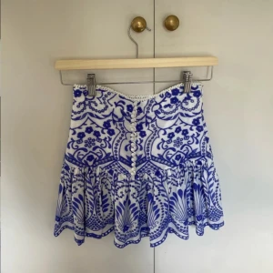 Vit och blå broderad kortkjol med knappar - Superfin kortkjol i vitt med blått broderat blommönster. Kjolen har volangkant, dekorativa knappar framtill och dragkedja i sidan. Tillverkad i bomull, perfekt för sommarens alla tillfällen. Söt och trendig design med fina detaljer.