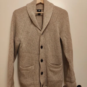 Beige stickad kofta från H&M - Mysig beige stickad kofta från H&M med sjalkrage, knappar framtill och två fickor. Tillverkad i en ullmix som håller dig varm. Perfekt för lager-på-lager och chill höst- eller vinterstil.