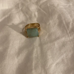 Guldfärgad ring med grön sten - Snygg guldfärgad ring med en stor, rektangulär ljusgrön sten i mitten. Ringen har en bred skena och en modern design som verkligen sticker ut. Perfekt accessoar för dig som vill ha något unikt och trendigt.