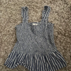 Blåvit randig linnetopp med volang - Säljer en söt blåvit randig linnetopp med smockad överdel och volang nedtill. Toppen har breda axelband med volangdetaljer och är perfekt för varma dagar. Materialet känns lätt och luftigt, och mönstret är klassiskt randigt.