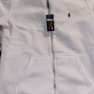 Vit hoodie från Polo Ralph Lauren - Snygg vit hoodie från Polo Ralph Lauren med dragkedja framtill, huva med snörning och klassisk logga på bröstet. Tröjan har känguruficka och är perfekt för en chill och stilren look. Pris kan diskuteras, den helt ny 