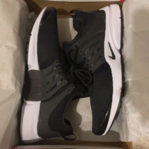 Nike Presto svarta sneakers - Nike Presto sneakers i svart med vit sula och diskreta Nike-loggor. Ovandelen är i mesh för bra ventilation och har snörning samt plastdetaljer på sidorna för extra stöd. Snygg, sportig design med rund tå och platt sula, perfekt för dig som gillar stilrena och bekväma skor.