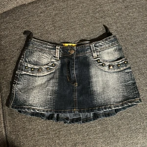 Lågmidjad kort jeanskjol - Snygg kort jeanskjol från Bleed i tvättad blå denim med slitningar och coola nitar på fickorna. Kjolen har volangkant nedtill, klassiska jeanssömmar och ett gult märke baktill. Perfekt för dig som gillar edgy streetstyle och detaljer.