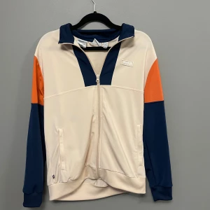 Adidas hoodie med färgblock - Snygg hoodie från Adidas i storlek M med dragkedja framtill. Tröjan har färgblock i beige, marinblått och orange, samt huva och två fickor. Materialet är mjuk polyester som känns skönt mot huden. Perfekt för dig som gillar sportig och modern stil.