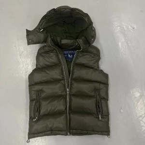 Olivgrön väst från Leon Marcél - Snygg olivgrön dunväst från Leon Marcél med avtagbar huva och två dragkedjeförsedda fickor. Västen har quiltad design och en rejäl dragkedja framtill. Perfekt för lager-på-lager och streetstyle. Märke syns på insidan.