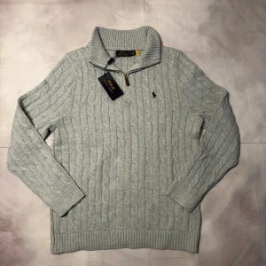 Ljusgrå stickad half zip från Polo Ralph Lauren - Säljer en ljusgrå stickad tröja från Polo Ralph Lauren med half zip och ribbad krage. Tröjan har ett klassiskt kabelstickat mönster, långärmade ärmar och den ikoniska Polo-loggan broderad på bröstet. Perfekt för dig som gillar stilren och chill look.