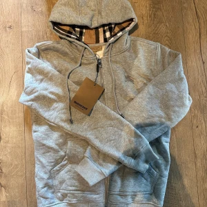 Grå hoodie från Burberry med dragkedja - Grå hoodie från Burberry med klassiskt rutigt foder i huvan och insidan. Tröjan har dragkedja, snörning vid huvan och broderad Burberry-logga på bröstet. Mjuk bomullskänsla och ribbade muddar. Perfekt för dig som gillar stilrena detaljer och ikoniskt mönster.