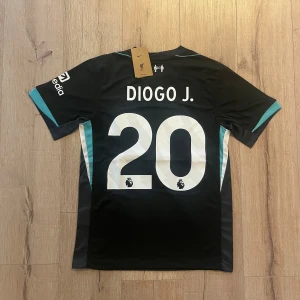 Liverpool FC svart bortatröja Nike - Liverpool FC bortatröja med Diogo J. och nummer 20 på ryggen. Svart med vita tryck och turkosa detaljer på ärmarna. Nike-logga och klubbmärke på bröstet, samt sponsortryck från Standard Chartered och Expedia. Tillverkad i lätt och ventilerande polyester.