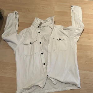 Vit overshirt med svarta knappar - Vit overshirt från Jack and Jones med svarta knappar och två bröstfickor med lock. Skjortan har klassisk krage och lång ärm, perfekt för lager-på-lager. Tillverkad i mjuk bomull för en avslappnad vibe. Har m vanliga tröjor så den passa lika bra till dd med 