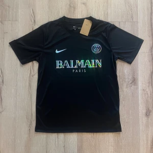 Svart PSG x Balmain fotbollströja Nike M - Svart fotbollströja från Nike i Dri-FIT-material, PSG x Balmain Paris edition. Kortärmad med rund hals och holografiskt Balmain-tryck på bröstet. PSG-logga och Nike Swoosh framtill. Perfekt för dig som gillar exklusiva samarbeten och sportig stil.