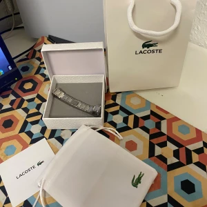 Silverfärgat armband från Lacoste - Stilrent armband från Lacoste i silverfärgad metall med länkdesign och diskret Lacoste-logga på mitten. Kommer i original ask och med tygpåse med broderad krokodil. Perfekt accessoar för en clean look.