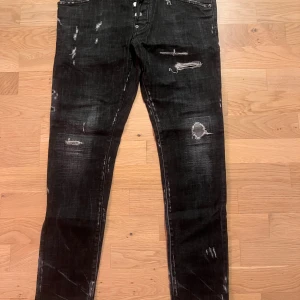 Svarta Dsquared2 Skater Jeans, stl 48 - Svarta Dsquared2 Skater Jeans med slitningar och trasiga detaljer för en edgy look. Jeansen har smal passform, klassisk femficksdesign och är tillverkade i bomull med en touch av stretch. Snygga kontrastsömmar och ikonisk 'ICON'-patch baktill.