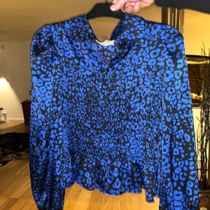 Blå leopardmönstrad skjorta - Snygg blå skjorta med svart leopardmönster och långärmad design. Skjortan har krage och smockad midja som ger en cool och trendig siluett. Perfekt för dig som vill sticka ut med ett djärvt mönster och färg. Materialet känns lätt och luftigt.