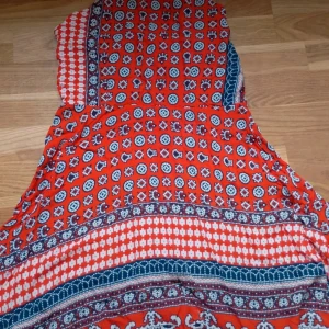 Röd mönstrad klänning - Färgstark klänning med rött, blått och vitt mönster i boho-stil. Klänningen har ärmlös design och ett mixat geometriskt och etniskt mönster. Perfekt för varma dagar och festivaler. Lätt och luftig känsla.