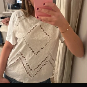 Vit blus med spets och hålmönster - Säljer en vit blus med korta ärmar och snygga spetsdetaljer. Blusen har geometriska hålmönster i form av rutor och zigzag, vilket ger en cool och unik look. Materialet är mjukt och har en lätt struktur med blommig spets. Perfekt för dig som gillar detaljer!