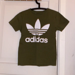 Olivgrön Adidas t-shirt med logga - Snygg olivgrön t-shirt från Adidas med stor vit trefoil-logga och text på bröstet. Klassisk rund hals och korta ärmar. Tillverkad i mjuk bomull som är skön mot huden. Perfekt för dig som gillar streetwear och sportig stil.