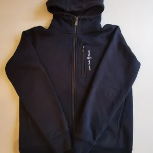 Marinblå hoodie från Sail Racing - Säljer en marinblå hoodie från Sail Racing med dragkedja framtill och logga på bröstet. Tröjan har huva, långa ärmar och en bröstficka med dragkedja. Perfekt för dig som gillar sportig och stilren look.