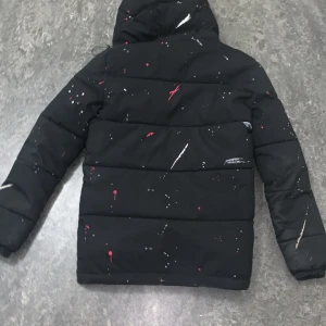 Svart pufferjacka Kings Will Dream - Svart pufferjacka från Kings Will Dream med coolt färgstänksmönster i rött och vitt. Jackan har huva med snörning, dragkedja framtill och långa ärmar. Perfekt för dig som vill sticka ut med streetstyle och hålla dig varm.