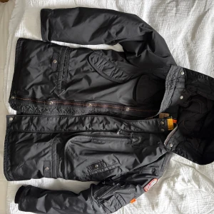 Svart Parajumpers pufferjacka med huva - Young small - Svart pufferjacka från Parajumpers med huva, flera fickor och patch på ärmen. Jackan har quiltad insida och snygga sömmar. Modellen är rymlig och har både knappar och dragkedja framtill. Observera att det är en junior jacka. Mycket gott skick.