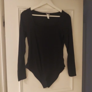 Svart långärmad bodysuit från H&M - Svart bodysuit från H&M med lång ärm och fyrkantig halsringning. Sitter tight och har en clean, minimalistisk look. Perfekt att styla med jeans eller kjol. Materialet är mjukt och stretchigt, troligen bomull med inslag av syntet för extra komfort.