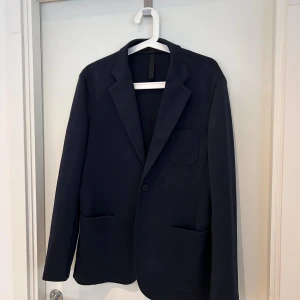 Mörkblå kavaj från J.Lindeberg - Snygg mörkblå kavaj från J.Lindeberg med klassisk krage och tre framfickor. Enkelknäppt design och stilren look som passar till många olika outfits. Materialet är mjukt och har en lätt struktur, vilket ger en modern känsla.