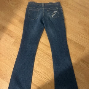 Von Dutch blå bootcut jeans - Snygga blå jeans från Von Dutch med diskret randigt mönster och klassisk bootcut-passform. Byxorna har fem fickor, bälteshällor och broderad Von Dutch-logga på bakfickan. Perfekta för dig som gillar en retro vibe och vill sticka ut lite extra.