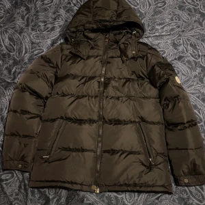 Svart Moncler pufferjacka med huva - Svart Moncler Montgenevre med huva och dragkedja framtill. STORLKE 3 (M). Jackan har två snedställda fickor med dragkedja, Moncler-logga på ärmen och quiltad design. Insidan har en cool serietidningsetikett och jackan är tillverkad i syntetmaterial med dunfyllning.