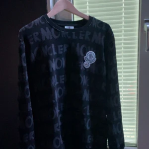 Svart långärmad  tröja - Svart långärmad tröja med coolt all-over logomönster och Moncler-patch på bröstet. Tröjan har rund hals och är tillverkad i mjukt material som känns skönt mot huden. Perfekt för dig som gillar streetwear och vill sticka ut med en snygg designerlogga.