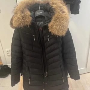 Svart pufferjacka Hollies med päls - Svart pufferjacka från Hollies med fluffig brun fuskpäls på huvan och leopardmönstrat foder. Jackan har flera dragkedjor framtill och snygga detaljer i orange på insidan. Perfekt för dig som vill ha en varm och cool vinterstil.