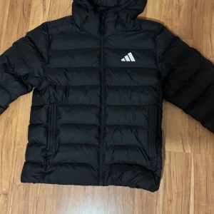 Svart pufferjacka från Adidas - Säljer en svart pufferjacka från Adidas med huva och dragkedja framtill. Jackan har ett stilrent Adidas-logga på bröstet och horisontella quiltade paneler. Regular fit och två sidofickor med dragkedja. Perfekt för kalla dagar.
