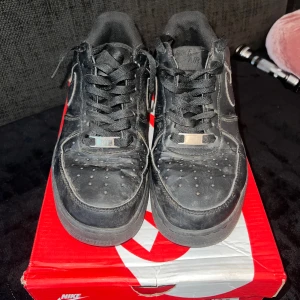 Svarta Nike Air Force 1 sneakers - Svarta Nike Air Force 1 sneakers med klassisk låg siluett och rund tå. Skorna har snörning, platt sula och är tillverkade i skinn med textilfoder. Swoosh-loggan syns på sidan och AIR-detalj på hälen. Perfekt för dig som gillar streetwear och tidlös stil.