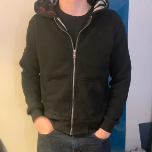 Svart Burberry hoodie med rutig huva och inredning  - Svart hoodie från Burberry med hel dragkedja och klassisk rutig insida i huvan med röda, vita och grå detaljer. Tröjan har huva, två fickor fram och ribbade muddar. Perfekt för dig som gillar stilrena streetwear-plagg med lyxig touch.