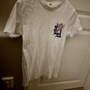 Vit t-shirt med lilla LA tryck - Säljer en vit t-shirt i storlek XS med ett LA tryck på bröstet i lila, där en bokstav kombineras med en liten ängel. T-shirten har rund hals och korta ärmar, perfekt för dig som gillar streetstyle och enkel design. Nästan aldrig andvänd. Andvänd 2-3 gånger